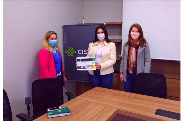 Na imagem, três mulheres de pé. A do meio segura um certificado.