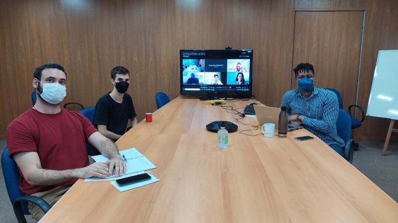 Três homens sentados em volta de mesa de reunião com uma televisão conectada em telechamada ao fundo.