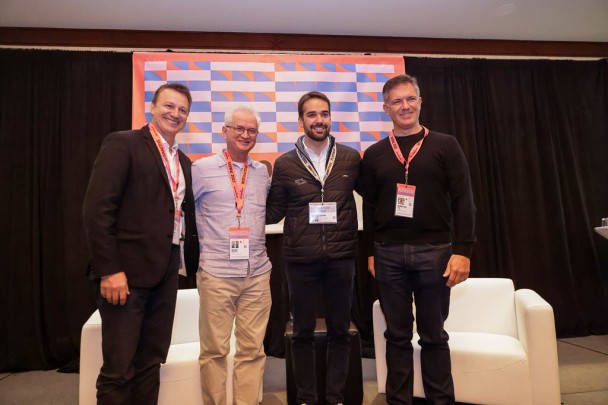 Quatro homens sorriem em frente a painel de conferência.