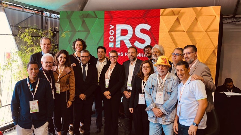 Pessoas reunidas atrás de um fundo com a logo do Estado do RS