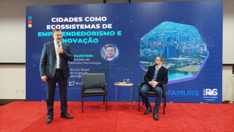Um homem de pé em frente a um painel e outro sentado.