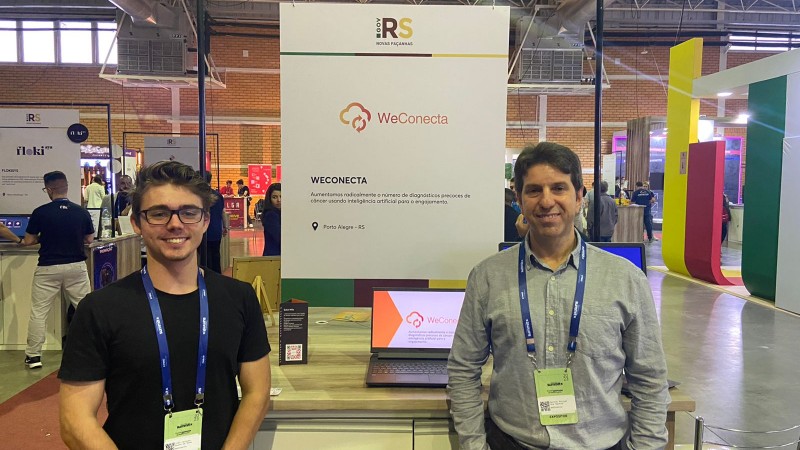 Dois homens brancos parados em frente a uma placa com o inscrito WeConecta, em uma feira de inovação.