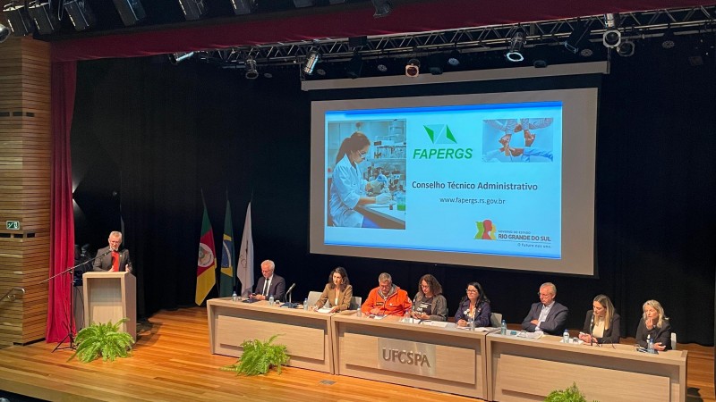 Foto da cerimônia, com presidente da Fapergs em pé no pulpito e demais autoridades sentadas à mesa durante apresentação do edital. 