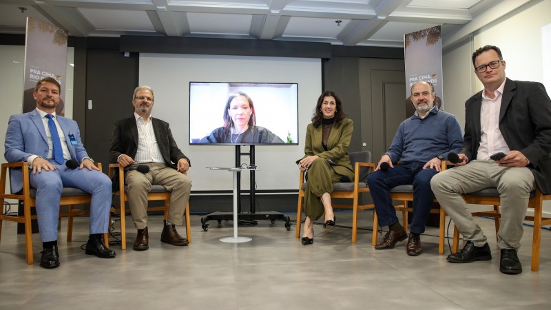 Todos os integrantes do painel estão posando para a foto. Os cinco integrantes presentes presencialmente estão sentados em cadeiras. Na ponta direita está o mediador, jornalista Rodrigo Lopes, ao seu lado, Rodrigo Sousa Costa - Presidente da Federasul e parceiro do Reconstrói RS, na sequência a secretária Simone Stülp, entre ela e o professor Jorge Audy há uma TV, na qual participa remotamente a professora e pesquisadora Taneha Kuzniecow Bacchin, por fim, na ponta esquerda está Arlei Romeiro - Presidente da Associação dos Empresários do 4º Distrito Vítimas da Enchente. 