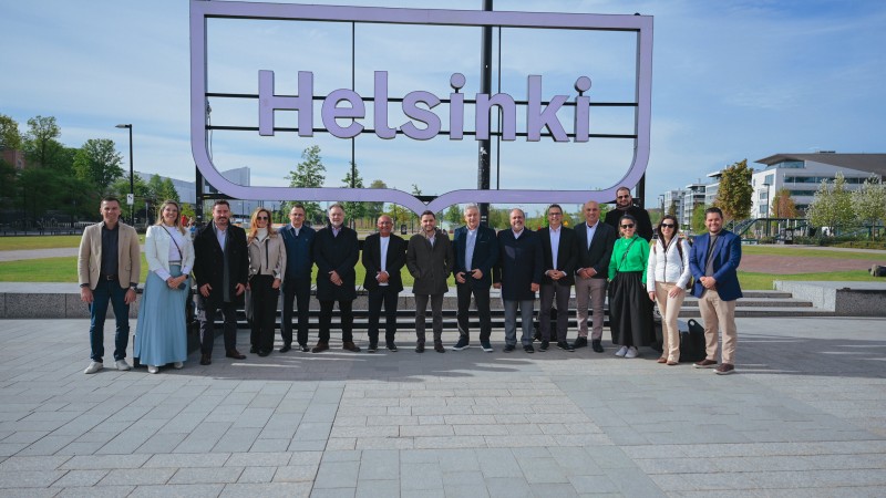 A foto mostra um grupo de 16 pessoas posando em frente a um grande letreiro que diz "Helsinki". Eles estão em uma área aberta com piso de pedra, cercada por grama e árvores, com prédios modernos ao fundo. A maioria está vestida de forma formal, com ternos e blazers, enquanto alguns usam roupas casuais, como jeans e uma jaqueta verde. O céu está claro, sugerindo um dia ensolarado.