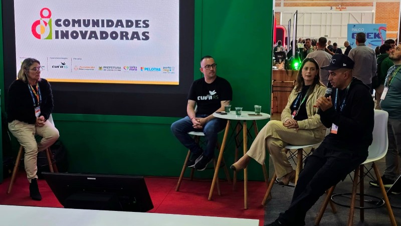 A imagem mostra quatro pessoas em um evento chamado "Comunidades Inovadoras". Elas estão sentadas em cadeiras e bancos ao redor de uma mesa redonda com copos d'água. Ao fundo, há uma tela com o logotipo do evento e nomes de parceiros como "OpenDemocracy", "PorAqui", "Fundação", "Prefeitura de Porto Alegre", "Siga", "Pelotas" e "Porto". O cenário é uma feira ou conferência, com outras pessoas e estandes visíveis. O chão é vermelho, e há uma parede verde ao lado da tela.