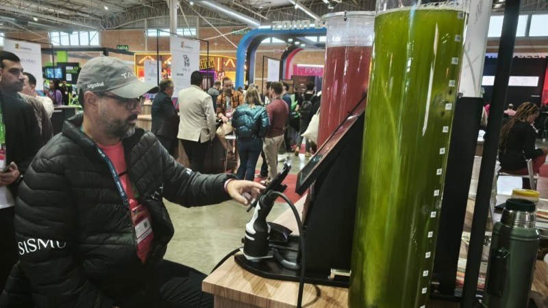 A imagem mostra um homem sentado em um evento ou feira, interagindo com uma máquina dispensadora de bebidas. Ele está usando uma jaqueta preta com a palavra "SISMU" no peito, uma camiseta vermelha por baixo e um crachá pendurado no pescoço. Ao seu lado, há um grande recipiente de vidro com uma bebida verde, possivelmente suco ou um shake, e outro recipiente com uma bebida vermelha. O ambiente é interno, com várias pessoas ao fundo, estandes e placas, sugerindo um ambiente de exposição ou convenção. Há também uma garrafa térmica verde sobre a mesa.