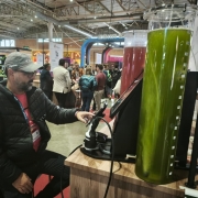 A imagem mostra um homem sentado em um evento ou feira, interagindo com uma máquina dispensadora de bebidas. Ele está usando uma jaqueta preta com a palavra "SISMU" no peito, uma camiseta vermelha por baixo e um crachá pendurado no pescoço. Ao seu lado, há um grande recipiente de vidro com uma bebida verde, possivelmente suco ou um shake, e outro recipiente com uma bebida vermelha. O ambiente é interno, com várias pessoas ao fundo, estandes e placas, sugerindo um ambiente de exposição ou convenção. Há também uma garrafa térmica verde sobre a mesa.