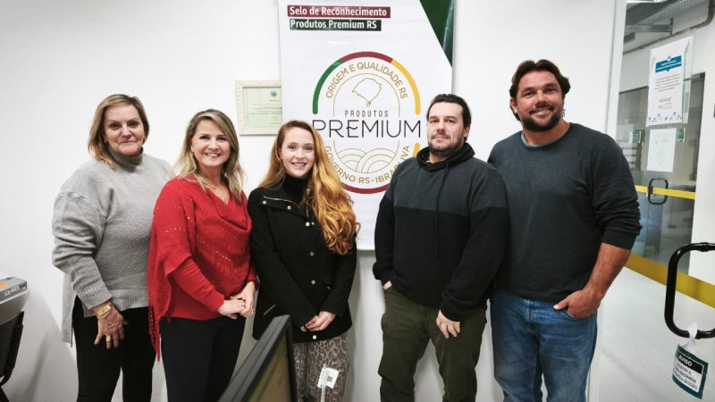 A imagem mostra cinco pessoas (três mulheres e dois homens) posando para uma foto em um ambiente interno, provavelmente um escritório ou instituição pública. Elas estão em frente a um banner que diz: "Produtos PREMIUM RS", com o selo "Origem e Qualidade RS - Governo RS - IBRAV" e a inscrição "Selo de Reconhecimento Produtos Premium RS" na parte superior. Esse selo parece estar relacionado ao reconhecimento da qualidade de produtos no estado do Rio Grande do Sul, Brasil.

As pessoas estão sorrindo e parecem estar participando de um momento comemorativo ou institucional. No ambiente, há uma mesa com objetos diversos (papéis, sacolas, pratos e copos), sugerindo uma confraternização ou reunião informal. Ao fundo, vê-se uma porta de vidro e parte de um corredor iluminado.

A atmosfera da foto é positiva, com todos bem vestidos e aparentando satisfação com o momento registrado.