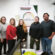 A imagem mostra cinco pessoas (três mulheres e dois homens) posando para uma foto em um ambiente interno, provavelmente um escritório ou instituição pública. Elas estão em frente a um banner que diz: "Produtos PREMIUM RS", com o selo "Origem e Qualidade RS - Governo RS - IBRAV" e a inscrição "Selo de Reconhecimento Produtos Premium RS" na parte superior. Esse selo parece estar relacionado ao reconhecimento da qualidade de produtos no estado do Rio Grande do Sul, Brasil.

As pessoas estão sorrindo e parecem estar participando de um momento comemorativo ou institucional. No ambiente, há uma mesa com objetos diversos (papéis, sacolas, pratos e copos), sugerindo uma confraternização ou reunião informal. Ao fundo, vê-se uma porta de vidro e parte de um corredor iluminado.

A atmosfera da foto é positiva, com todos bem vestidos e aparentando satisfação com o momento registrado.
