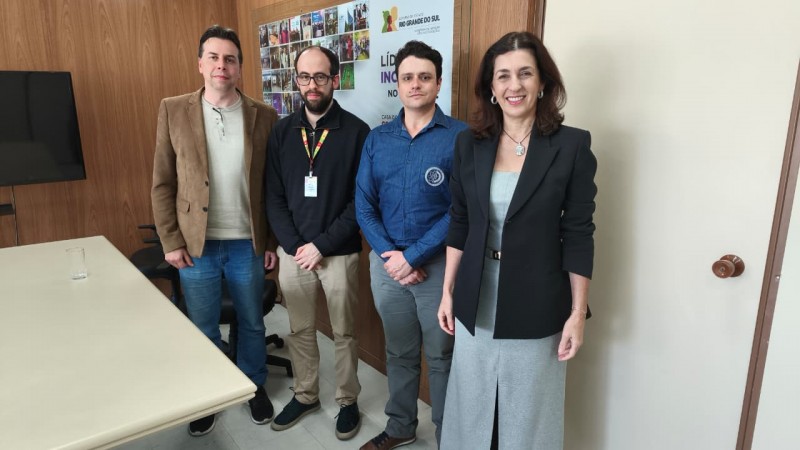 A imagem mostra quatro pessoas posando em um ambiente de escritório com paredes de madeira e um cartaz ao fundo que menciona "Liderança Inovadora" e "Rio Grande do Sul". Há uma mesa à esquerda com um copo d'água e uma cadeira, além de uma televisão montada na parede. As pessoas estão vestidas de forma casual a semiformal, com uma mulher à direita usando um blazer e saia, e os homens usando camisas e calças variadas. O ambiente parece ser uma sala de reuniões ou escritório institucional.