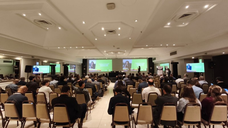 A imagem mostra um evento ou confer&ecirc;ncia realizado em um audit&oacute;rio fechado, possivelmente em um centro de conven&ccedil;&otilde;es ou sal&atilde;o de hotel. O espa&ccedil;o est&aacute; bem iluminado, com fileiras de cadeiras ocupadas por participantes, todos voltados para a frente, onde h&aacute; tr&ecirc;s grandes telas de proje&ccedil;&atilde;o.
As telas exibem slides com fundo verde e a imagem de uma pessoa falando, indicando que est&aacute; ocorrendo uma palestra ou apresenta&ccedil;&atilde;o. O p&uacute;blico parece atento, sugerindo um ambiente profissional e organizado, t&iacute;pico de semin&aacute;rios, f&oacute;runs ou encontros corporativos.