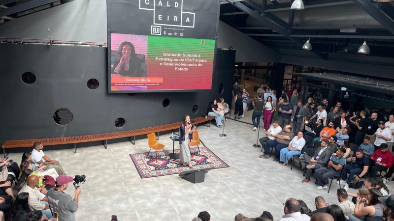 Evento em espa&ccedil;o moderno com p&uacute;blico atento, palco central com microfone e tel&atilde;o ao fundo exibindo o tema &ldquo;Gramado Summit e Estrat&eacute;gias do C&T para o Desenvolvimento do Estado&rdquo;. Ambiente marcado por networking e inova&ccedil;&atilde;o.