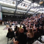 Auditório moderno com teto envidraçado, lotado de pessoas assistindo a uma apresentação no palco central. Um grande telão exibe informações do evento, enquanto o público ocupa cadeiras e arquibancadas em clima de networking.