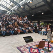 Auditório moderno com arquibancadas cheias de participantes atentos, enquanto uma pessoa apresenta no palco com microfone. À frente, há duas cadeiras sobre um tapete colorido e um monitor exibindo conteúdo do evento.