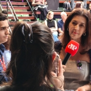 Pessoa sendo entrevistada em um ambiente de evento, segurando um microfone vermelho com logotipo de veículo de imprensa. Ao fundo, há arquibancadas com público sentado e escadas, indicando clima de cobertura jornalística.