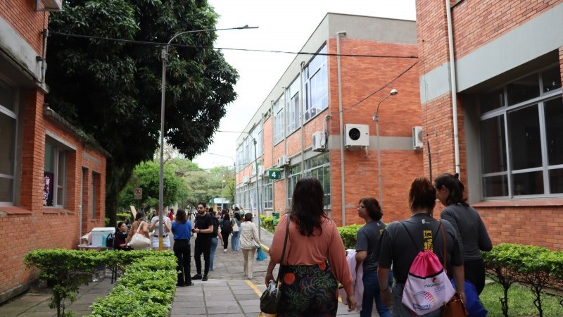 A imagem mostra uma passarela entre dois pr&eacute;dios de tijolos com janelas grandes, provavelmente em um campus universit&aacute;rio. H&aacute; pessoas caminhando, &aacute;rvores e arbustos bem cuidados ao longo do caminho, criando um ambiente organizado e movimentado.