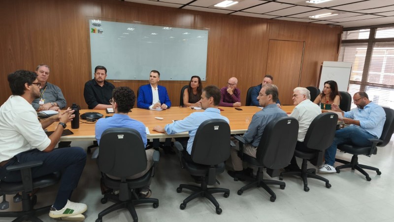 Reuni&atilde;o em sala corporativa com v&aacute;rias pessoas sentadas ao redor de uma mesa retangular, discutindo assuntos. Ao fundo, h&aacute; um quadro branco e, &agrave; direita, uma janela ampla com entrada de luz natural.
