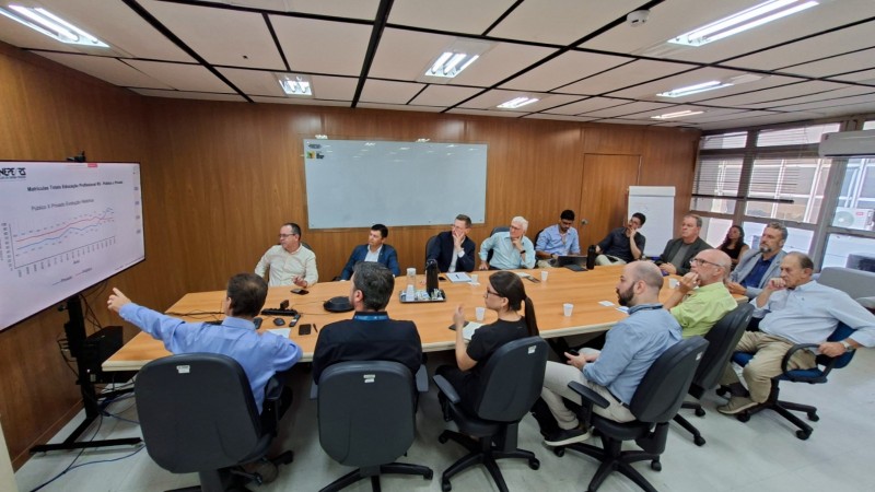 Grupo de pessoas numa sala de reuni&otilde;es, junto &agrave; mesa, de frente para uma tela de tv, com um homem apontando para a tela.