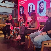 Num palco, quatro pessoas, três mulheres e um homem, estão lado a lado com um painel eletrônico atrás.