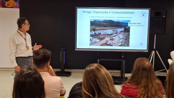 Homem ao lado de uma tela de tv, apresenta imagens para a plateia.