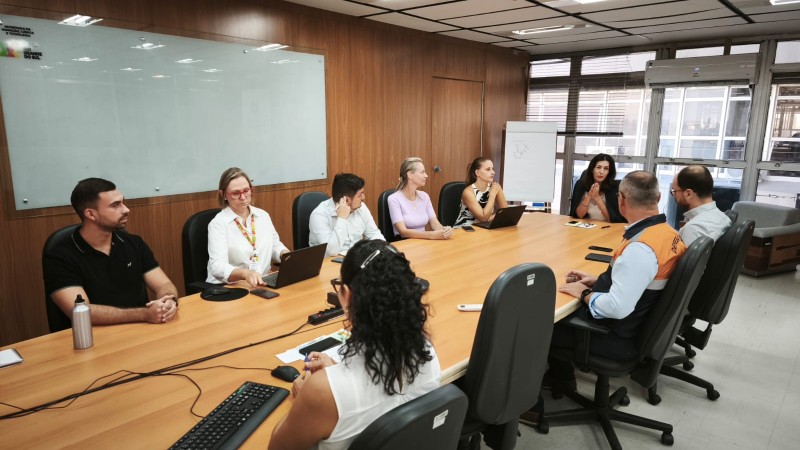 A imagem mostra uma reuni&atilde;o em uma sala ampla com mesa retangular, onde v&aacute;rias pessoas est&atilde;o sentadas e utilizando computadores e cadernos. O ambiente possui parede de madeira, quadro branco e janelas grandes ao fundo. A disposi&ccedil;&atilde;o sugere um encontro de trabalho ou discuss&atilde;o t&eacute;cnica em andamento.