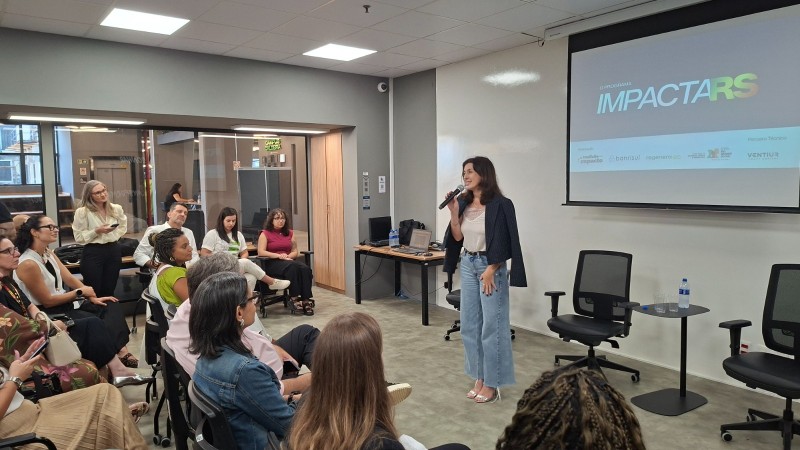 mulher com microfone na m&atilde;o de frente para plateia e tel&atilde;o no fundo.