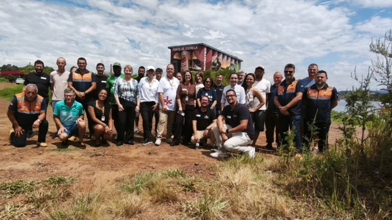 A imagem mostra um grupo reunido em &aacute;rea externa, posicionado para uma foto coletiva em frente a um pr&eacute;dio com mural colorido. O cen&aacute;rio ao redor inclui vegeta&ccedil;&atilde;o baixa, c&eacute;u parcialmente nublado e um corpo d&rsquo;&aacute;gua ao fundo.