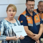 A imagem mostra três pessoas vestindo uniformes de Defesa Civil ao lado de outra pessoa que segura uma folha de papel com o texto “GESTÃO DE RISCO” impresso. O grupo está em um ambiente interno, com cadeiras claras ao fundo e paredes em tons neutros.