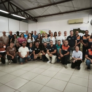 A imagem mostra um grande grupo reunido em uma sala, organizado em duas fileiras para uma foto coletiva após a atividade. Ao fundo, mapas e materiais de apoio indicam um ambiente voltado à capacitação e gestão de riscos.