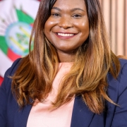 Uma mulher sorridente, de cabelos longos castanhos com mechas claras, vestindo blazer escuro e blusa clara, posa diante de um fundo institucional com bandeira.