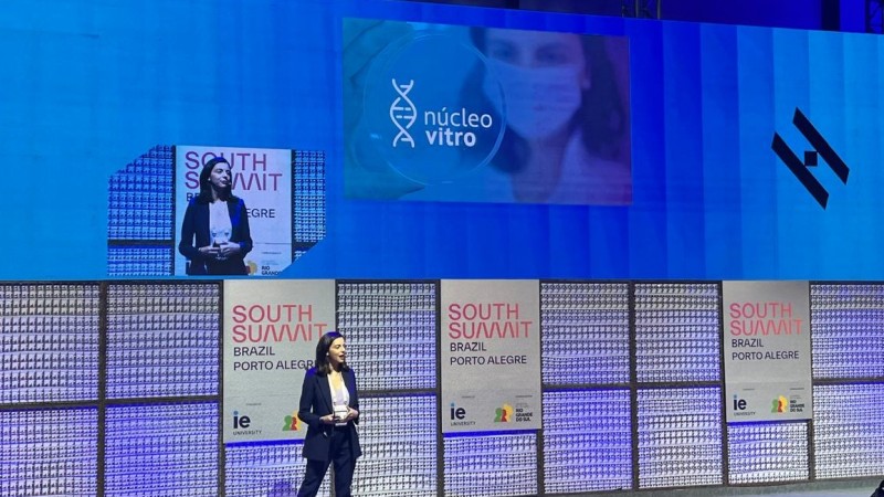 Uma pessoa apresenta no palco do South Summit Brazil, em Porto Alegre, diante de um grande painel azul com a marca &ldquo;n&uacute;cleo vitro&rdquo; projetada ao fundo.
O cen&aacute;rio &eacute; composto por tel&otilde;es, ilumina&ccedil;&atilde;o c&ecirc;nica e pain&eacute;is do evento, refor&ccedil;ando o contexto de inova&ccedil;&atilde;o e startups.