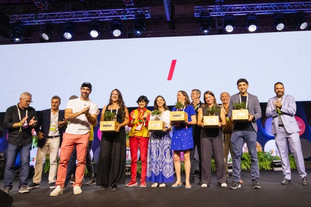 Um grupo de participantes ocupa o palco de um evento, posando lado a lado enquanto recebem prêmios e são aplaudidos.
Ao fundo, um grande telão claro e a iluminação cênica reforçam o clima de celebração e reconhecimento coletivo.