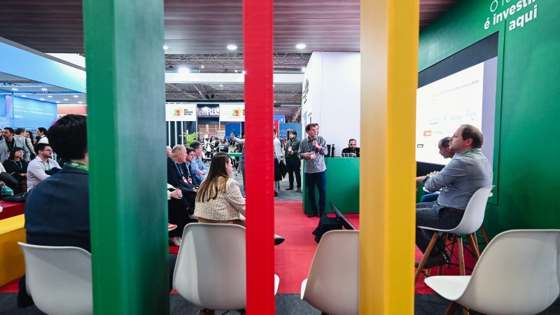A imagem mostra um ambiente de feira ou confer&ecirc;ncia. Em primeiro plano, quatro faixas verticais coloridas &mdash; verde, vermelho, amarelo e verde &mdash; dividem a cena. Ao fundo, h&aacute; v&aacute;rias pessoas sentadas assistindo a uma apresenta&ccedil;&atilde;o. Um palestrante est&aacute; de p&eacute; pr&oacute;ximo a uma tela grande, onde aparecem logotipos e textos relacionados a neg&oacute;cios e investimentos. O espa&ccedil;o tem estandes e marcas vis&iacute;veis, refor&ccedil;ando o clima de evento corporativo. Na parede verde atr&aacute;s do palestrante, l&ecirc;-se a frase em portugu&ecirc;s: &ldquo;O futuro &eacute; investir aqui.&rdquo;
