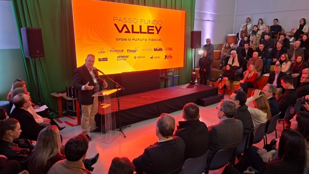Palestrante fala em um palco diante de plateia, com tel&atilde;o laranja ao fundo exibindo &ldquo;Passo Fundo Valley &ndash; onde o futuro nasce&rdquo; e logos de apoiadores.
Audit&oacute;rio moderno com p&uacute;blico sentado em cadeiras e arquibancada, ilumina&ccedil;&atilde;o quente e foco no evento de inova&ccedil;&atilde;o.