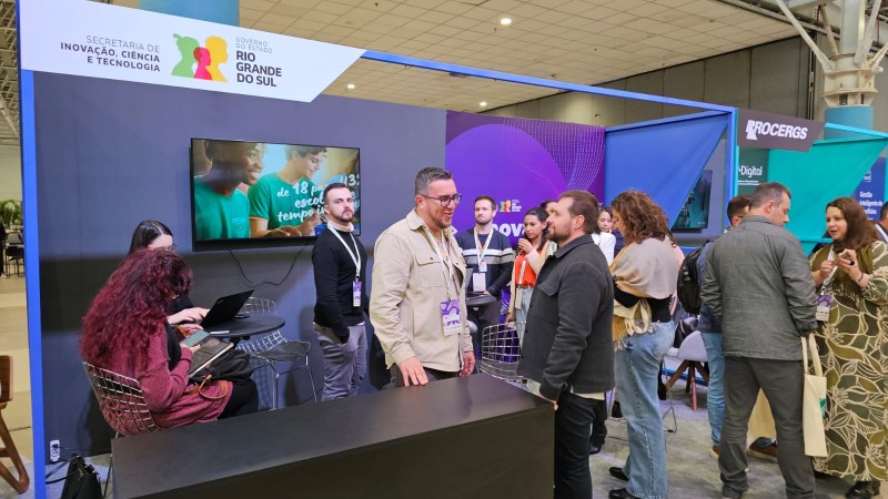 Estande da Secretaria de Inova&ccedil;&atilde;o, Ci&ecirc;ncia e Tecnologia do Rio Grande do Sul em um evento, com v&aacute;rias pessoas conversando e circulando pelo espa&ccedil;o.
Pain&eacute;is com logos institucionais e telas ao fundo, al&eacute;m de um balc&atilde;o de atendimento onde participantes interagem e usam computadores e celulares.