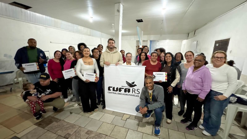 Descri&ccedil;&atilde;o da imagem: Grupo de aproximadamente 25 pessoas posa para uma foto coletiva em uma sala iluminada, com paredes brancas e piso de cer&acirc;mica. Ao centro, est&aacute; uma faixa com o logotipo da CUFA RS (Central &Uacute;nica das Favelas). Diversos participantes seguram certificados de conclus&atilde;o de curso. As pessoas est&atilde;o dispostas em p&eacute; e algumas agachadas, sorrindo para a c&acirc;mera. Na parte frontal, &agrave; esquerda, um homem est&aacute; sentado segurando uma crian&ccedil;a no colo; &agrave; direita, um homem est&aacute; agachado, sorrindo. A imagem registra um momento de integra&ccedil;&atilde;o e celebra&ccedil;&atilde;o da forma&ccedil;&atilde;o.
