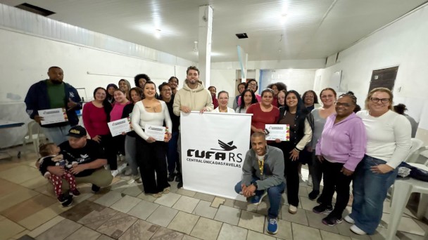 Descri&ccedil;&atilde;o da imagem: Grupo de aproximadamente 25 pessoas posa para uma foto coletiva em uma sala iluminada, com paredes brancas e piso de cer&acirc;mica. Ao centro, est&aacute; uma faixa com o logotipo da CUFA RS (Central &Uacute;nica das Favelas). Diversos participantes seguram certificados de conclus&atilde;o de curso. As pessoas est&atilde;o dispostas em p&eacute; e algumas agachadas, sorrindo para a c&acirc;mera. Na parte frontal, &agrave; esquerda, um homem est&aacute; sentado segurando uma crian&ccedil;a no colo; &agrave; direita, um homem est&aacute; agachado, sorrindo. A imagem registra um momento de integra&ccedil;&atilde;o e celebra&ccedil;&atilde;o da forma&ccedil;&atilde;o.
