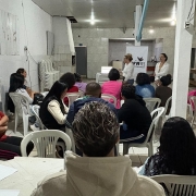 Participantes estão sentados em cadeiras de plástico brancas, organizadas em fileiras, acompanhando uma apresentação em uma sala comunitária. Ao fundo, duas mulheres estão em pé diante do grupo, conduzindo a atividade. Uma delas usa blazer branco e calça cinza; a outra veste roupas claras. Atrás delas, há uma faixa com o logotipo da CUFA RS e um computador sobre uma mesa. Os participantes estão voltados para as apresentadoras, e alguns têm cadernos e folhas sobre as mesas. À direita, há uma mesa com materiais e objetos. A imagem registra um momento de formação e troca de conhecimentos durante a oficina.