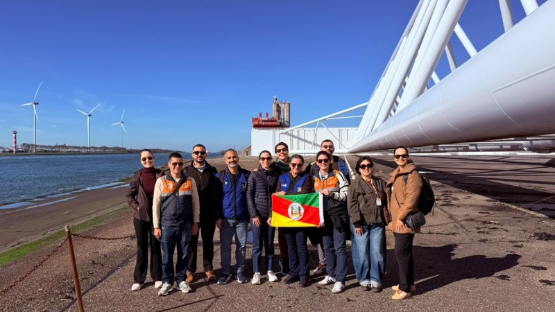 Grupo de pessoas posa &agrave; beira de um canal, segurando a bandeira do Rio Grande do Sul. Ao fundo, grandes turbinas e&oacute;licas e uma ampla estrutura industrial branca se destacam sob c&eacute;u azul sem nuvens.