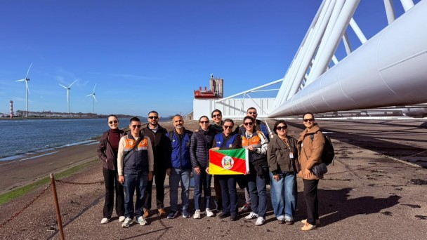 Grupo de pessoas posa &agrave; beira de um canal, segurando a bandeira do Rio Grande do Sul. Ao fundo, grandes turbinas e&oacute;licas e uma ampla estrutura industrial branca se destacam sob c&eacute;u azul sem nuvens.