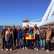 Grupo de pessoas posa &agrave; beira de um canal, segurando a bandeira do Rio Grande do Sul. Ao fundo, grandes turbinas e&oacute;licas e uma ampla estrutura industrial branca se destacam sob c&eacute;u azul sem nuvens.